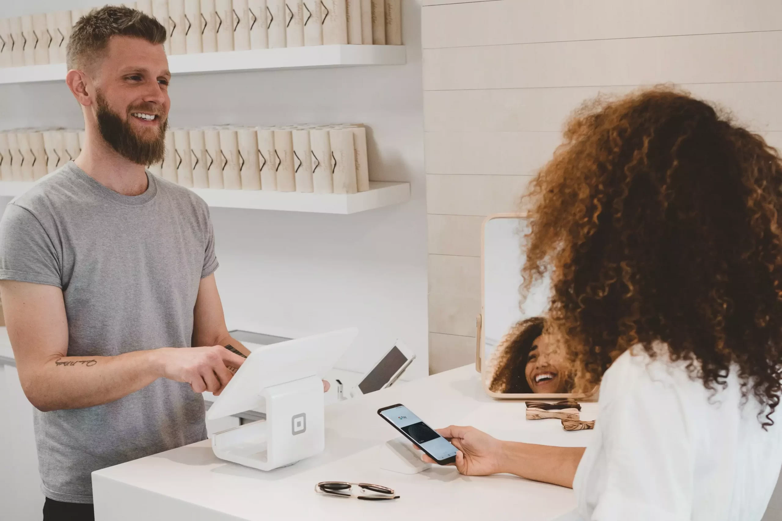 A girl is holdin a phone in white shirt and a guy in front of her holding a pad and smiling
